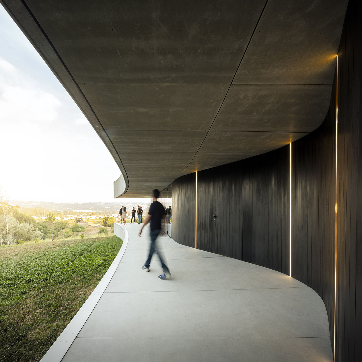 Cabo de Vila House, Paredes: covered walkway along a dark curved wall, sheltered by the cantilevered concrete slab