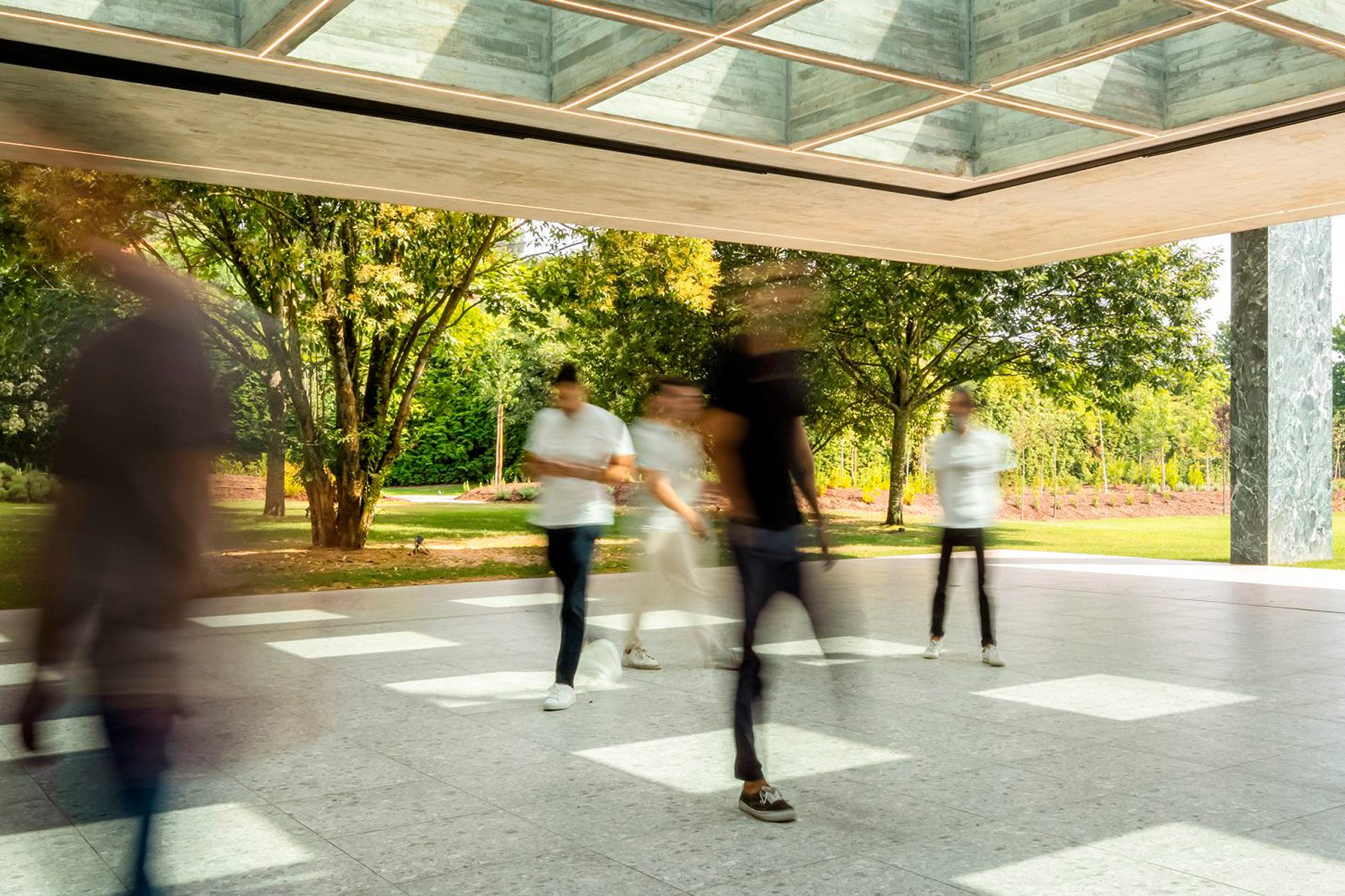 Pavilion in the Garden, Paredes: people crossing the open covered area beneath the glazed roof grid, blurred in motion
