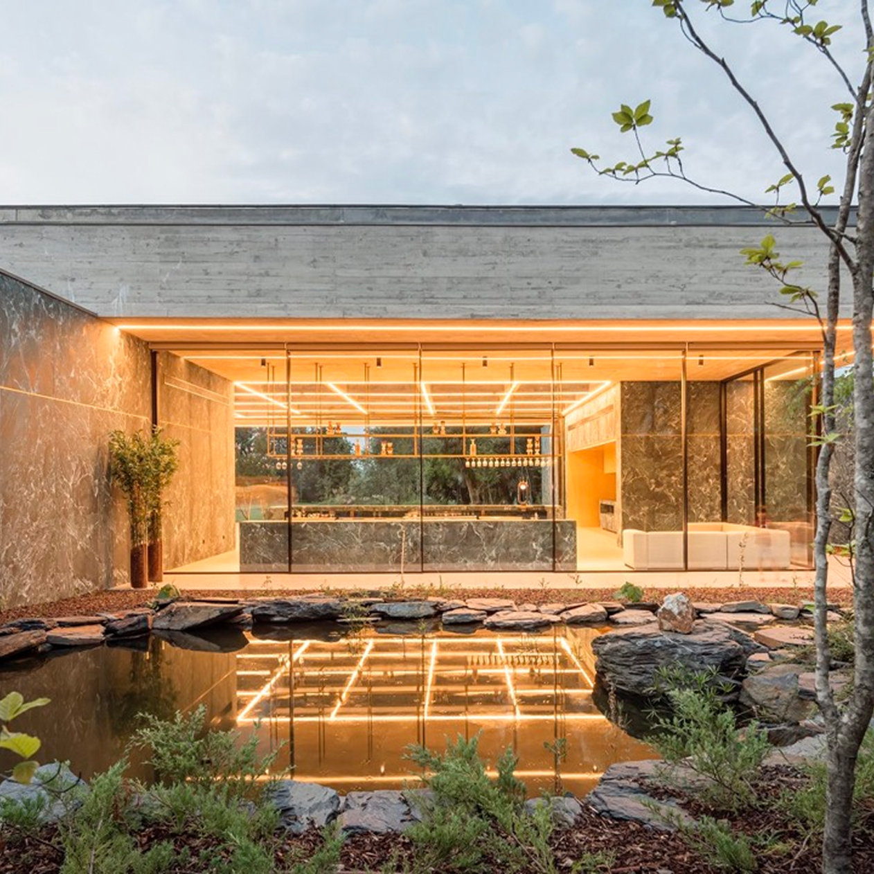 Pavilion in the Garden, Paredes: front view of the glazed interior with its lit ceiling reflected in the pond and stones