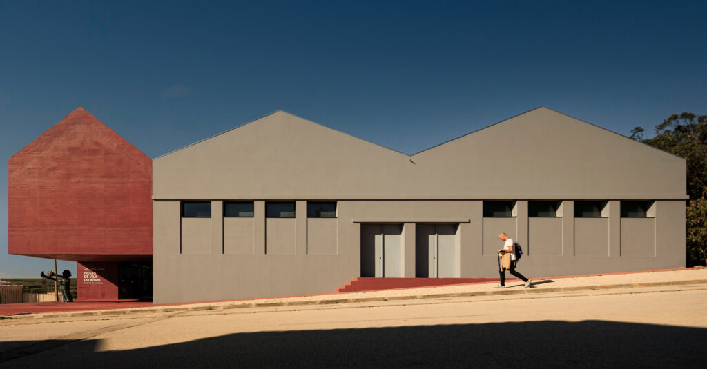 Street view of the Vila do Bispo Museum in the Algarve: the red concrete volume beside the grey gabled naves of the old grain stores