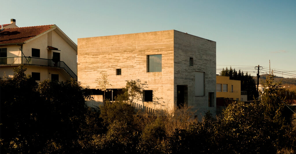 SV House in Sobrado, a concrete block with small scattered openings beside traditional pitched-roof houses
