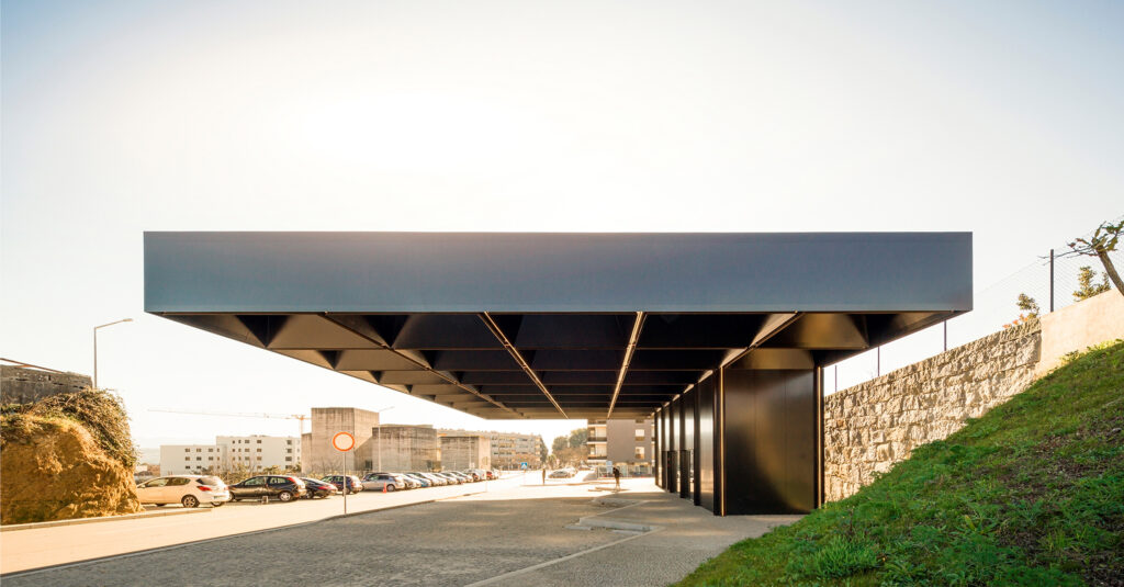 Lousada Intermodal Hub: a dark cantilevered roof hovering over the road beside a grassy embankment and stone wall