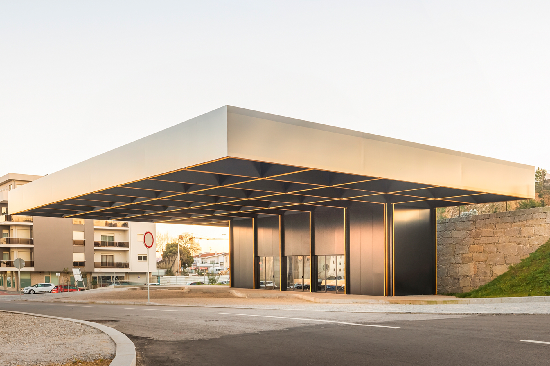 Lousada Intermodal Hub: street-level view of the roof spanning over the pavement, with a granite retaining wall and grass behind it