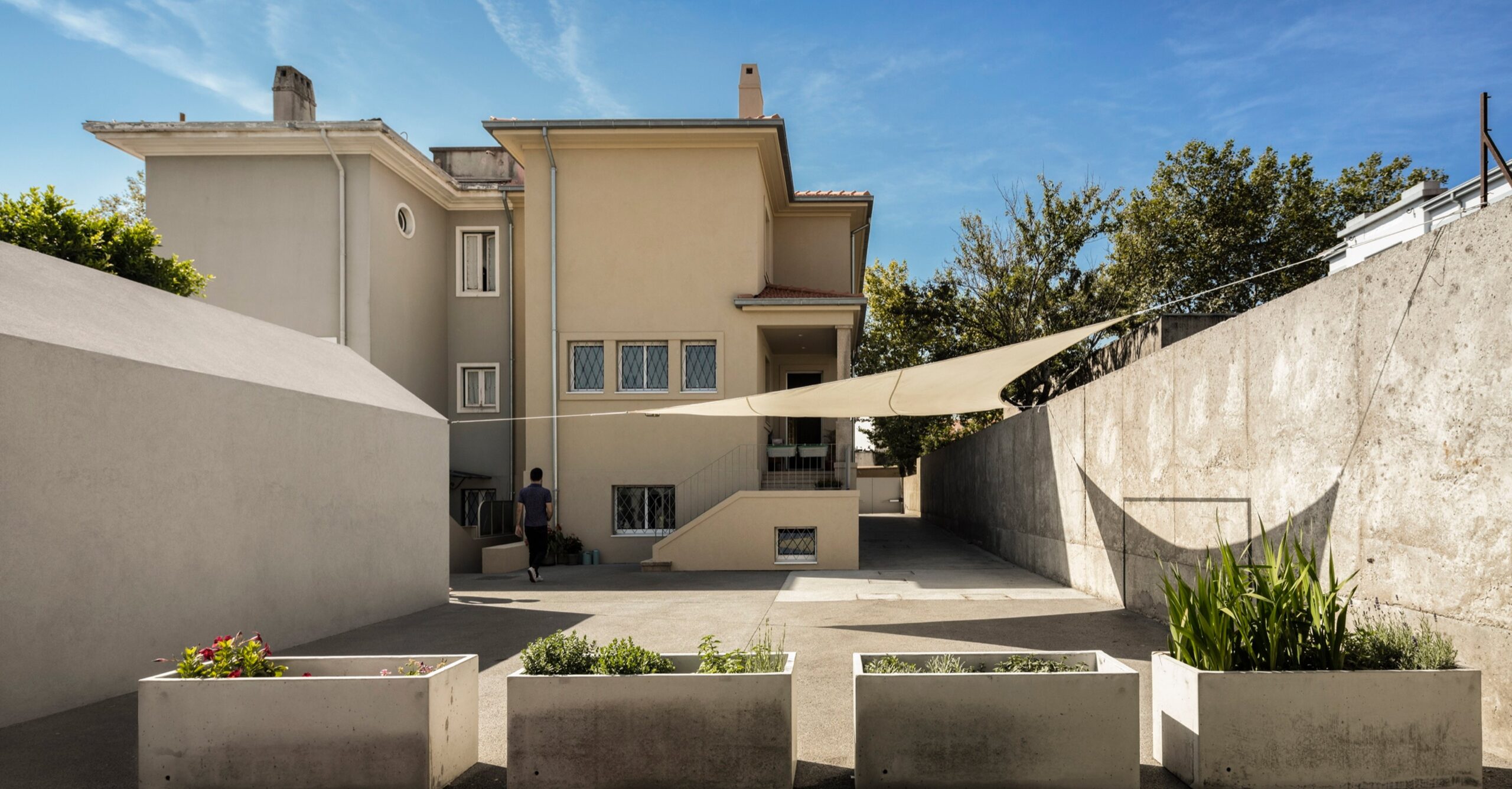 Combatentes House in Porto, the rear facade of the 1960s semi-detached house seen from the backyard, with shade sails and concrete planters